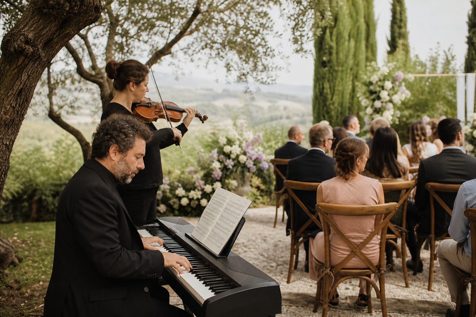 Pianist and violinist performing under trees beside seated guests at an outdoor wedding ceremony.
