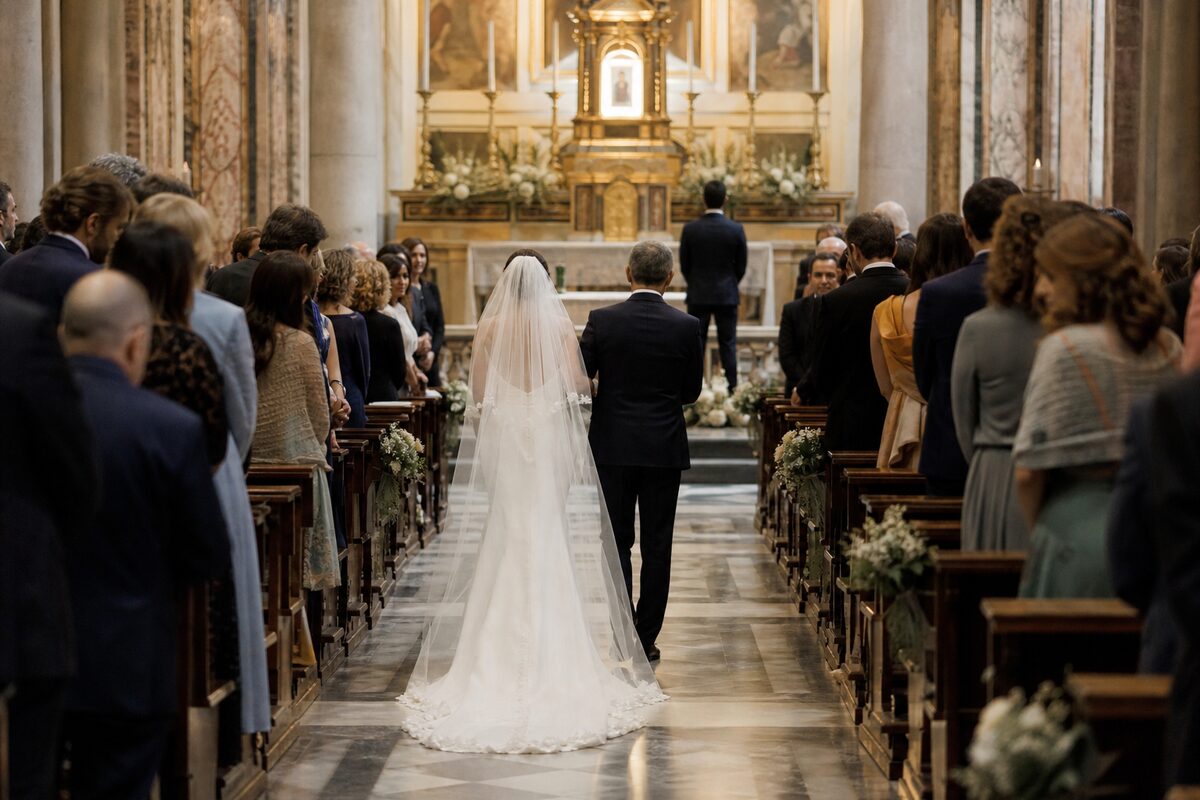 Rear view of a bride and her companion walking through standing guests toward a groom at the church altar.